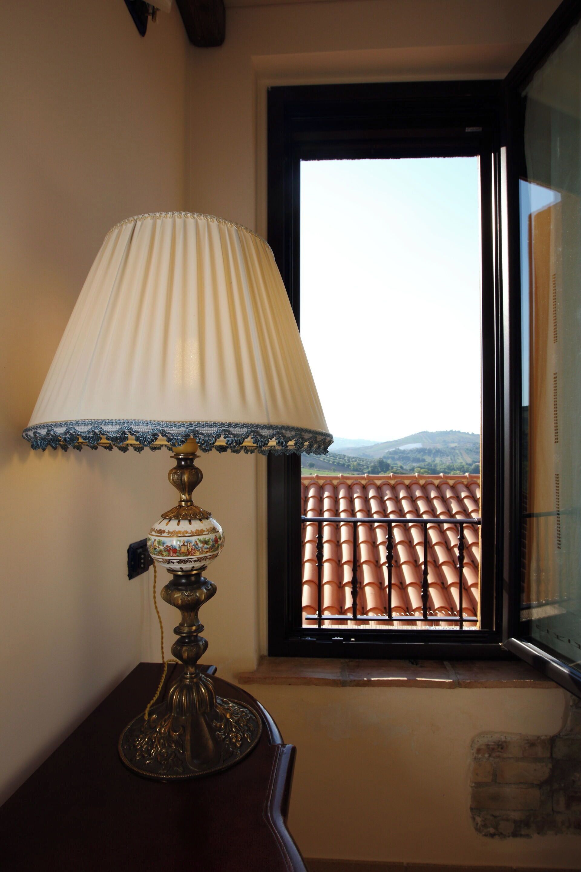 Une lampe avec un abat-jour décoratif, posée sur une table près d’une fenêtre donnant sur un toit recouvert de tuiles et sur des collines lointaines.