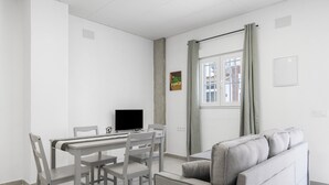 Ein modernes Wohnzimmer mit grauem Sofa, einem Esstisch mit Stühlen, einem Flachbildfernseher und einem Fenster mit Vorhängen.