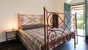 Ein Bett mit Metallgestell und einem karierten Bettbezug, ein Nachttisch mit einer Lampe und Blick auf Grün hinter einer offenen Tür.