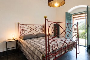A bed with a metal frame and a plaid bedspread, a bedside table with a lamp, and a view of greenery through an open door. - Villa Chiaramonte Gulfi (Chiaramonte Gulfi)