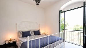 Ein Schlafzimmer mit einem Bett, Nachttischlampen, einem Balkon mit Geländer und Blick ins Grüne.