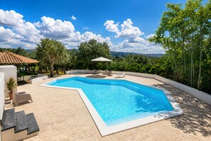 En swimmingpool med hvid kant, omgivet af en beige terrasse, og en pergola med en hvid parasol.