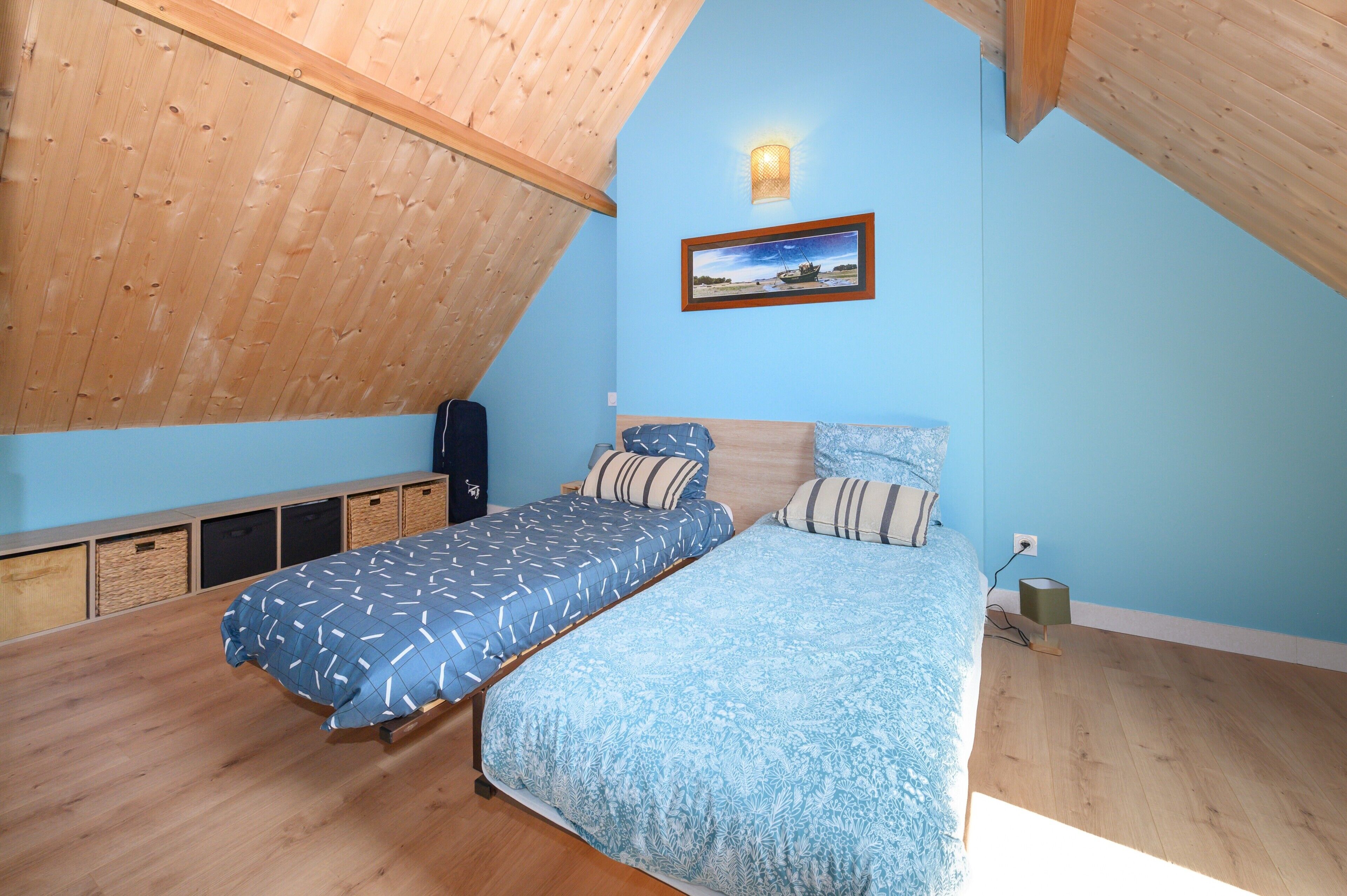 Een slaapkamer met twee bedden, houten vloeren, een schuin plafond, een lijst met een afbeelding aan de muur en een bedlampje.