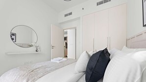 Una cama bien hecha con almohadas blancas y negras, un espejo redondo en la pared y un armario blanco con la puerta abierta hacia un pasillo.