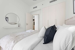 Ein ordentlich gemachtes Bett mit weißen und schwarzen Kissen, ein runder Spiegel an der Wand und ein weißer Kleiderschrank mit offener Tür zu einem Flur.