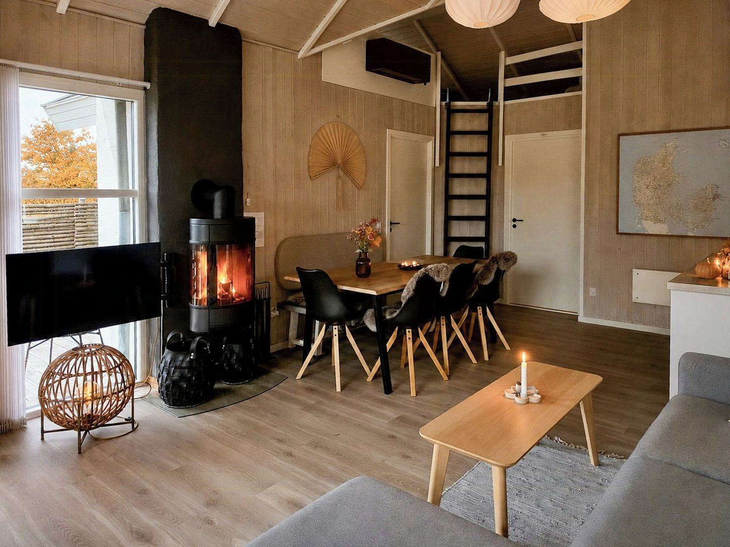 En hyggelig stue med en brændeovn, et spiseområde med stole, et fjernsyn og et stort vindue.