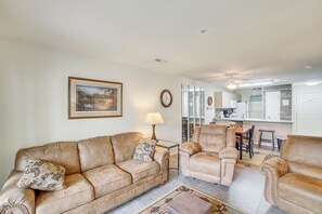 Ein Wohnzimmer mit einer Couch, Sesseln, einem Couchtisch, einer Lampe, einem gerahmten Bild und einer Küche im Hintergrund.