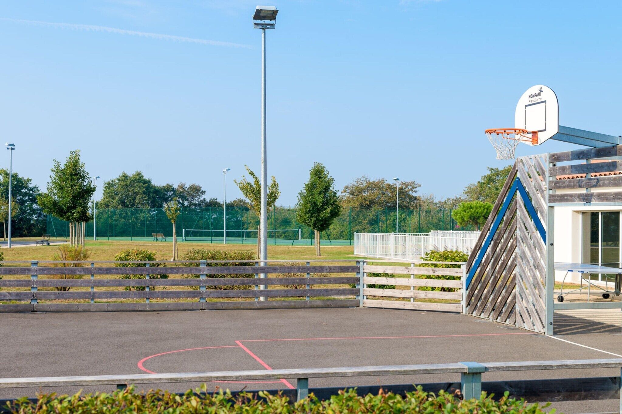 An outdoor basketball court with a wooden fence, a basketball hoop, and a building with a blue and white striped awning.