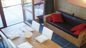 Un comedor con una mesa de madera dispuesta para cuatro personas, un sofá con cojines rojos, y vistas al exterior a través de un ventanal.