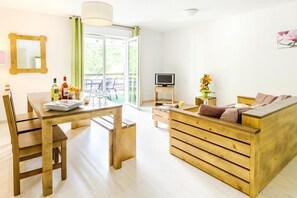 A room with a wooden dining table, chairs, a wooden sofa, a TV on a stand, and a window with a view of greenery.