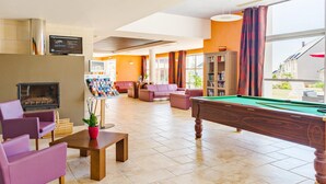 Ein Billardtisch, Sofas, ein Kamin und ein Fernseher in einem hellen Raum mit großen Fenstern.