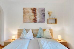 Una cama doble con almohadas estampadas, un cabecero de madera, mesitas de noche con lámparas, un cuadro abstracto en la pared y un pequeño estante de madera con plantas secas.