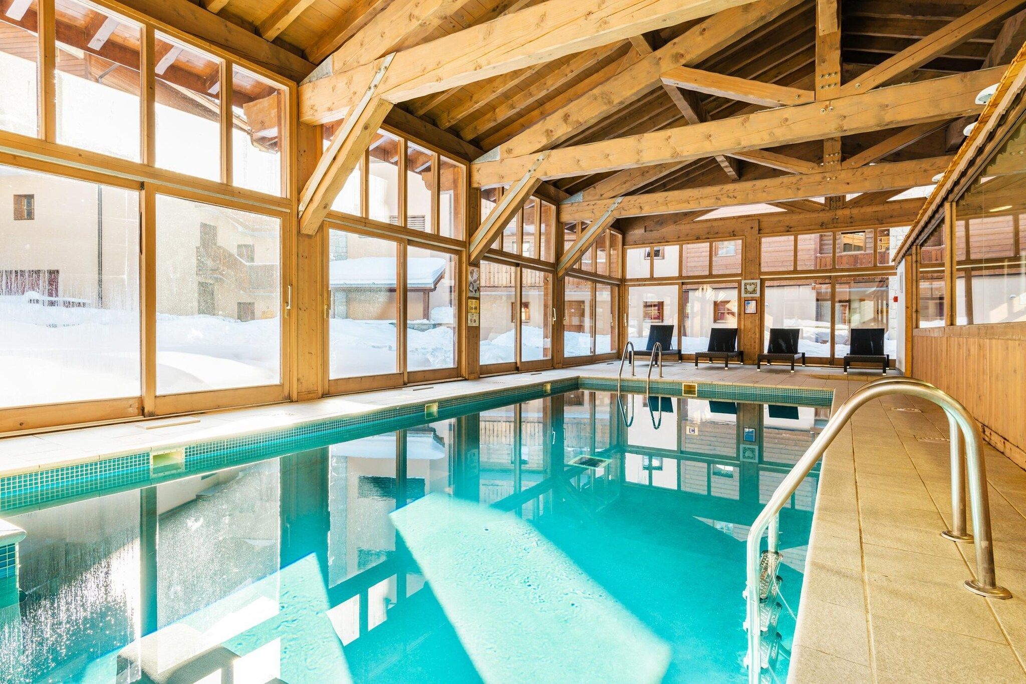 An indoor swimming pool with wooden beams, glass walls, and a clear view of the snowy outdoors.