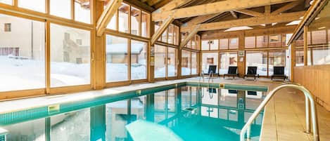 An indoor swimming pool with wooden beams, glass walls, and a clear view of the snowy outdoors.