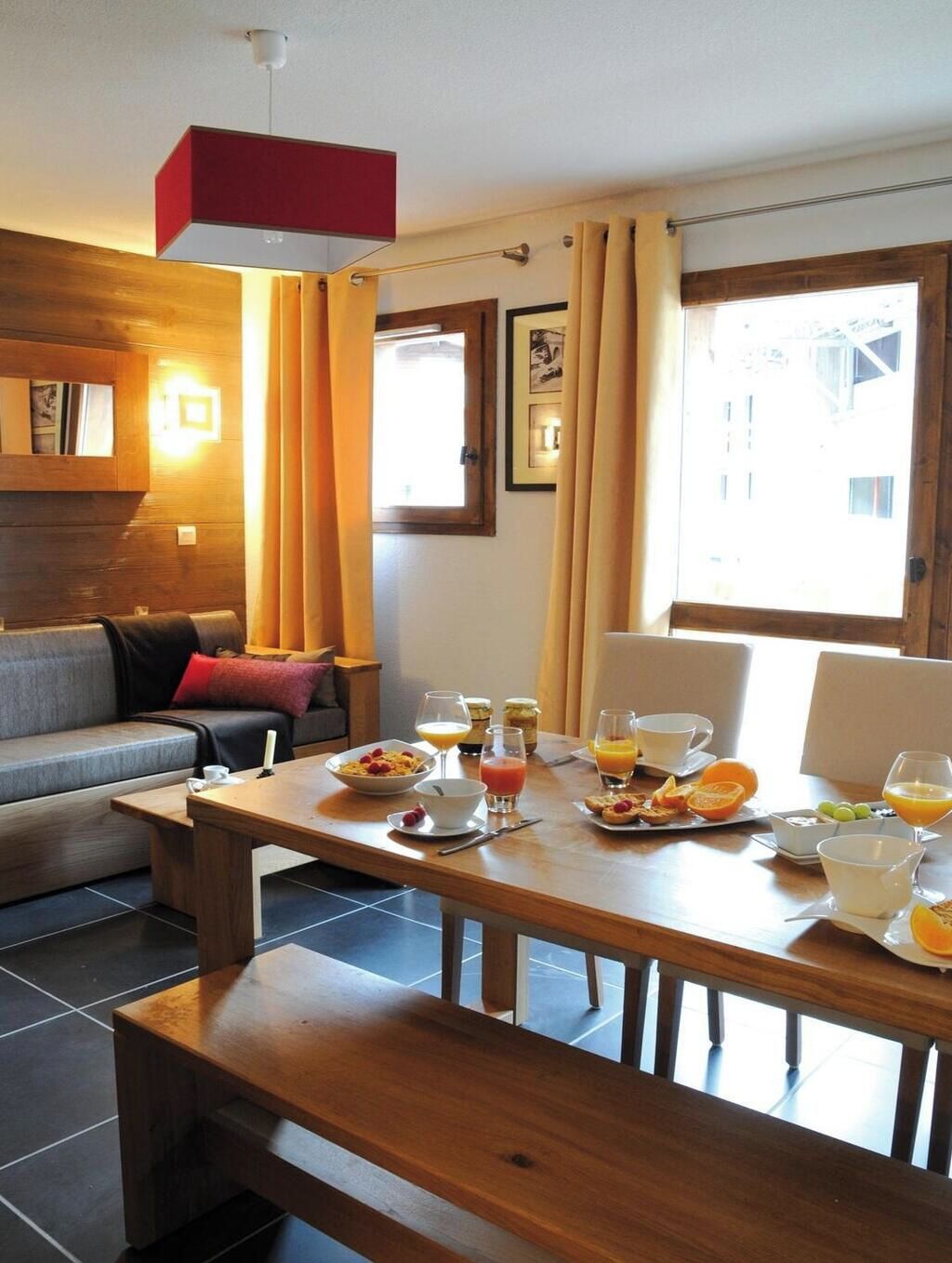 A dining area with a wooden table set for breakfast, a sofa, and a window with curtains.