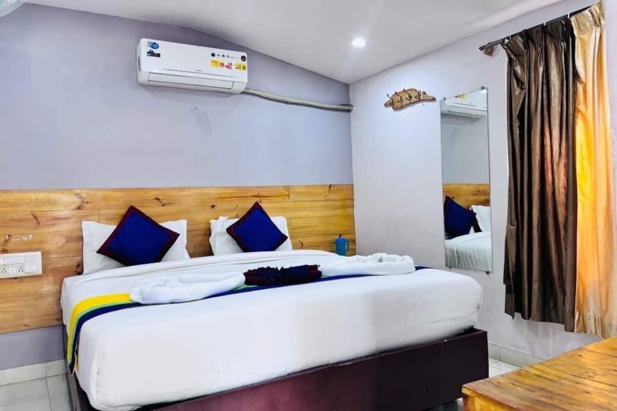 A hotel room with a bed, a wooden headboard, a wall-mounted air conditioner, a mirror, and a curtain.