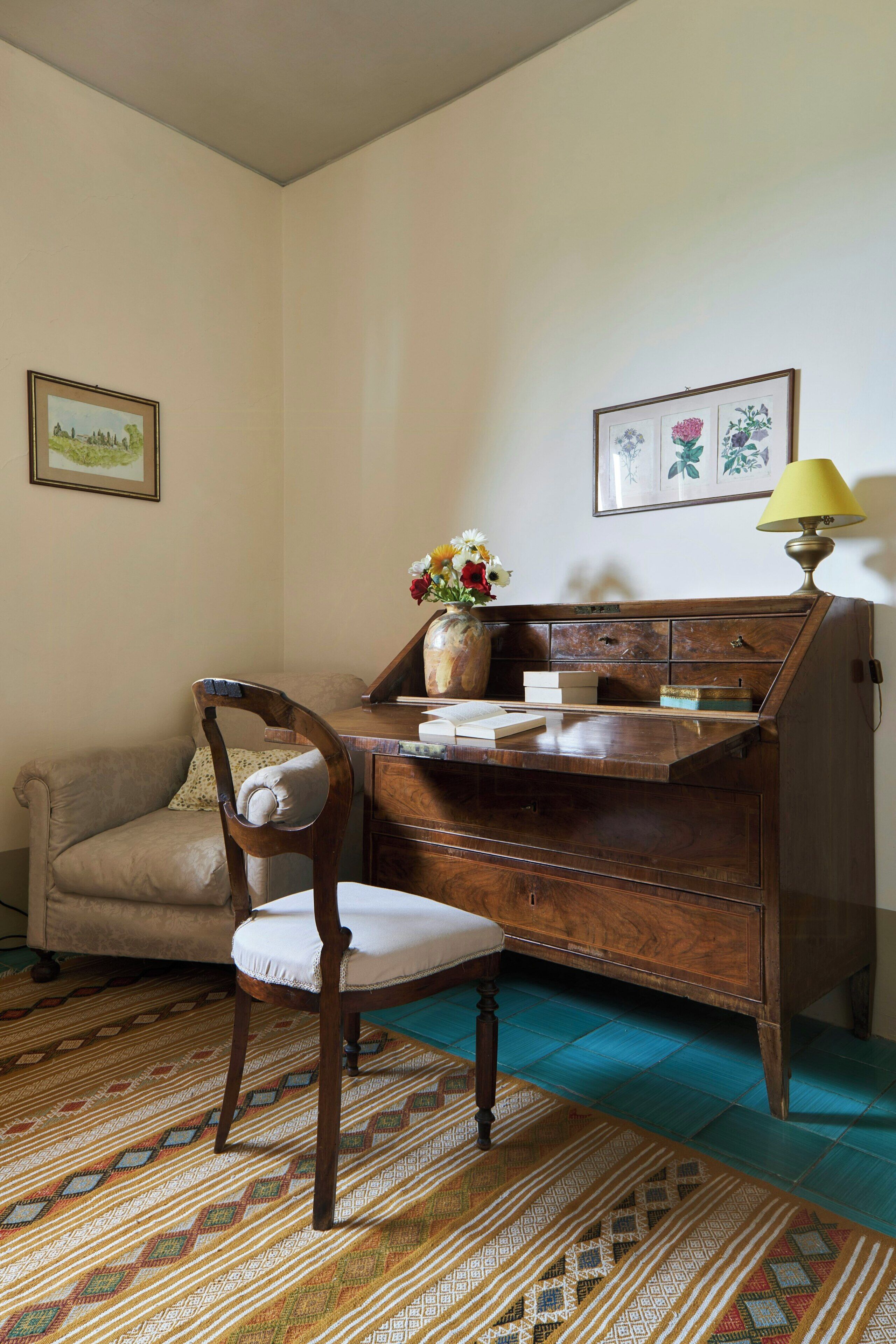 A room with a wooden desk, a chair, a sofa, a patterned rug, and a lamp.