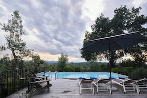 Un terrazzo in legno con poltrona lounge e piscina, circondato da alberi e un cielo nuvoloso.