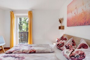 A bedroom with a bed, floral bedding, a view of greenery, and a painting on the wall.