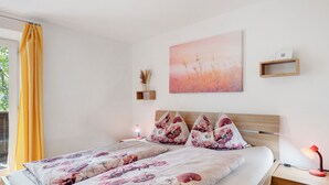 A double bed with floral bedding, a bedside table with a lamp, a nightstand with a lamp, a wall-mounted shelf with a vase, and a painting on the wall.