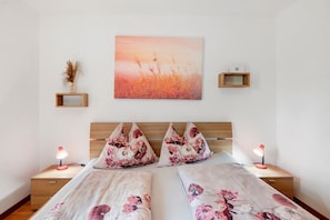 Una camera da letto con una testiera in legno, due comodini con lampade, un letto con biancheria da letto a motivi floreali, un quadro appeso al muro e una piccola mensola in legno con un oggetto decorativo.