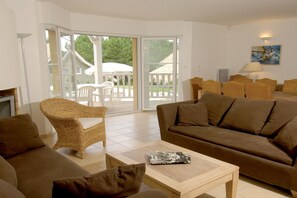 Ein Wohnzimmer mit einer Couch, Sesseln, einem Couchtisch und einer Glastür, die zu einem Außenbereich mit einem weißen Sonnenschirm und Stühlen führt.