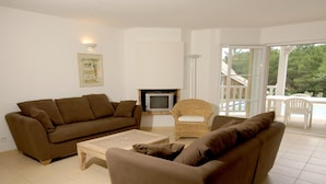 A living room with a brown sofa, a wicker chair, a small table, a fireplace, and a view of a balcony with outdoor furniture. - Villa for 6 People with Swimming Pool (Lacanau)