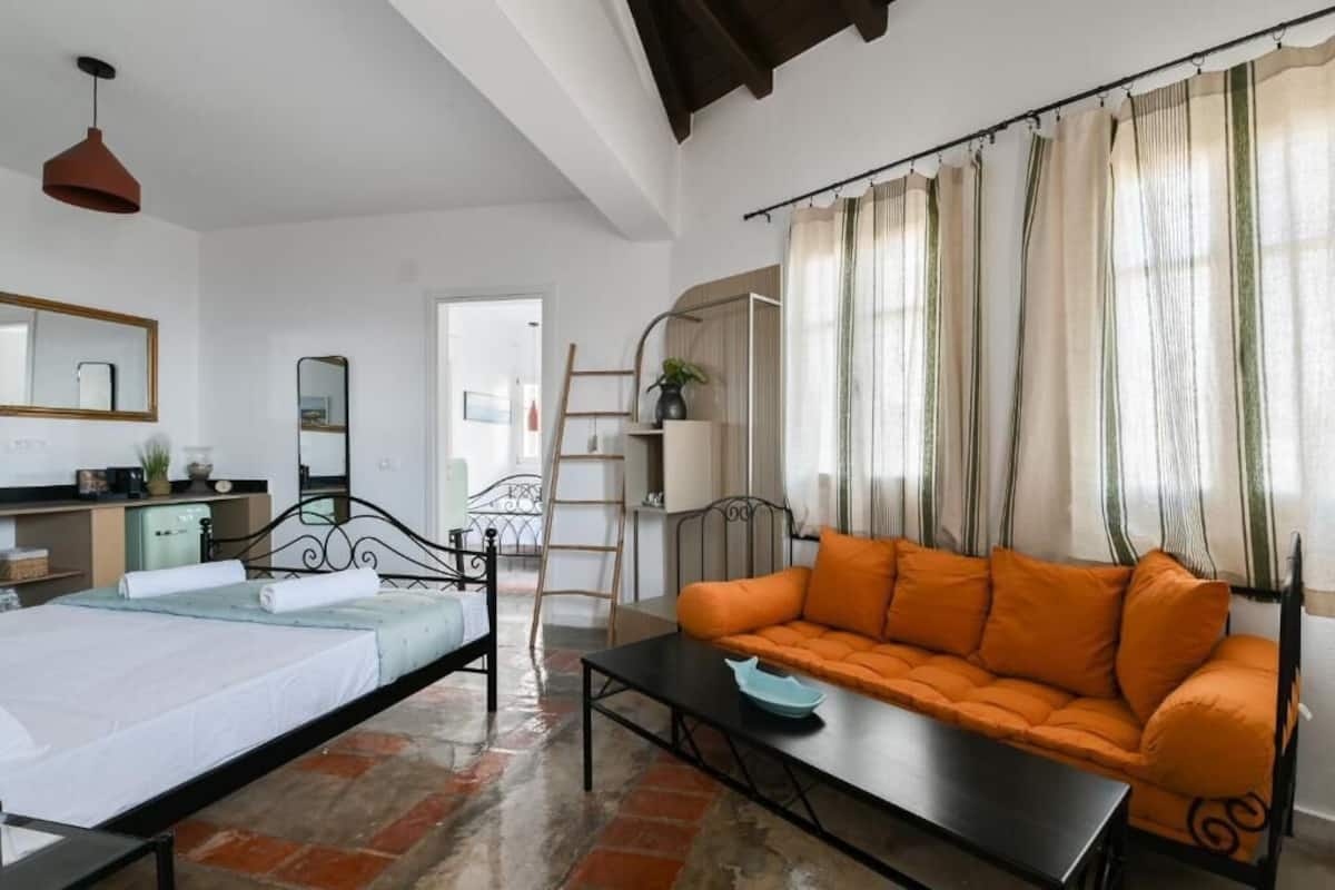 Una habitación con cama, sofá, mesa y espejo. Hay una escalera y una planta en la esquina.