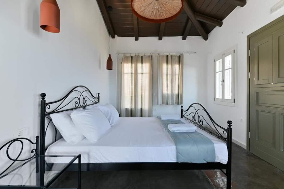 Una cama doble con estructura metálica, una mesita de vidrio, techo de madera y una puerta verde.