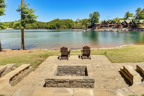 Un patio junto al lago con sillas Adirondack, un pozo de fuego y una cabaña al fondo.