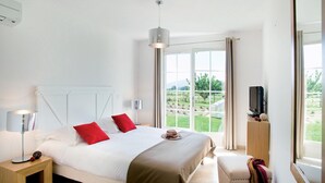 Un dormitorio con cama, televisión, ventana con cortinas y vistas al verdor exterior.