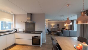 A modern kitchen with a central island, a dining area with a wooden table and chairs, and a living space with a sofa and a lamp. - Modern, luxurious and very comfortable holiday home with sauna, jacuzzi, BBQ Green Egg and trampoline (Colijnsplaat)