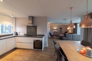 A modern kitchen with a central island, a dining area with a wooden table and chairs, and a living space with a sofa and a lamp. - Modern, luxurious and very comfortable holiday home with sauna, jacuzzi, BBQ Green Egg and trampoline (Colijnsplaat)
