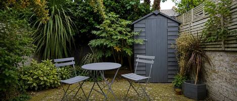 Un petit espace extérieur aménagé en patio, avec une table et deux chaises, entouré d’une végétation luxuriante et d’un abri en bois.