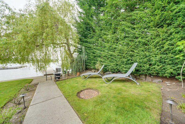 A serene outdoor area with a pathway leading to a seating area by a lake, surrounded by lush greenery and a weeping willow tree. - Executive Lake Washington Stay with Dock Access (Kirkland)