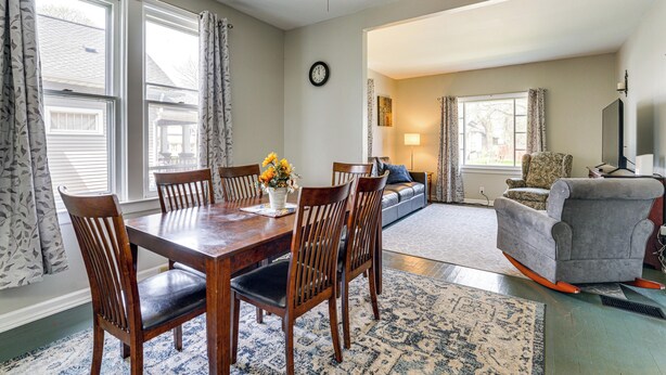 A dining area with a wooden table and chairs, a clock on the wall, and a living room with a sofa and a television. - Gateway to Dtwn Port Huron & Lake! Cozy Home (Port Huron)