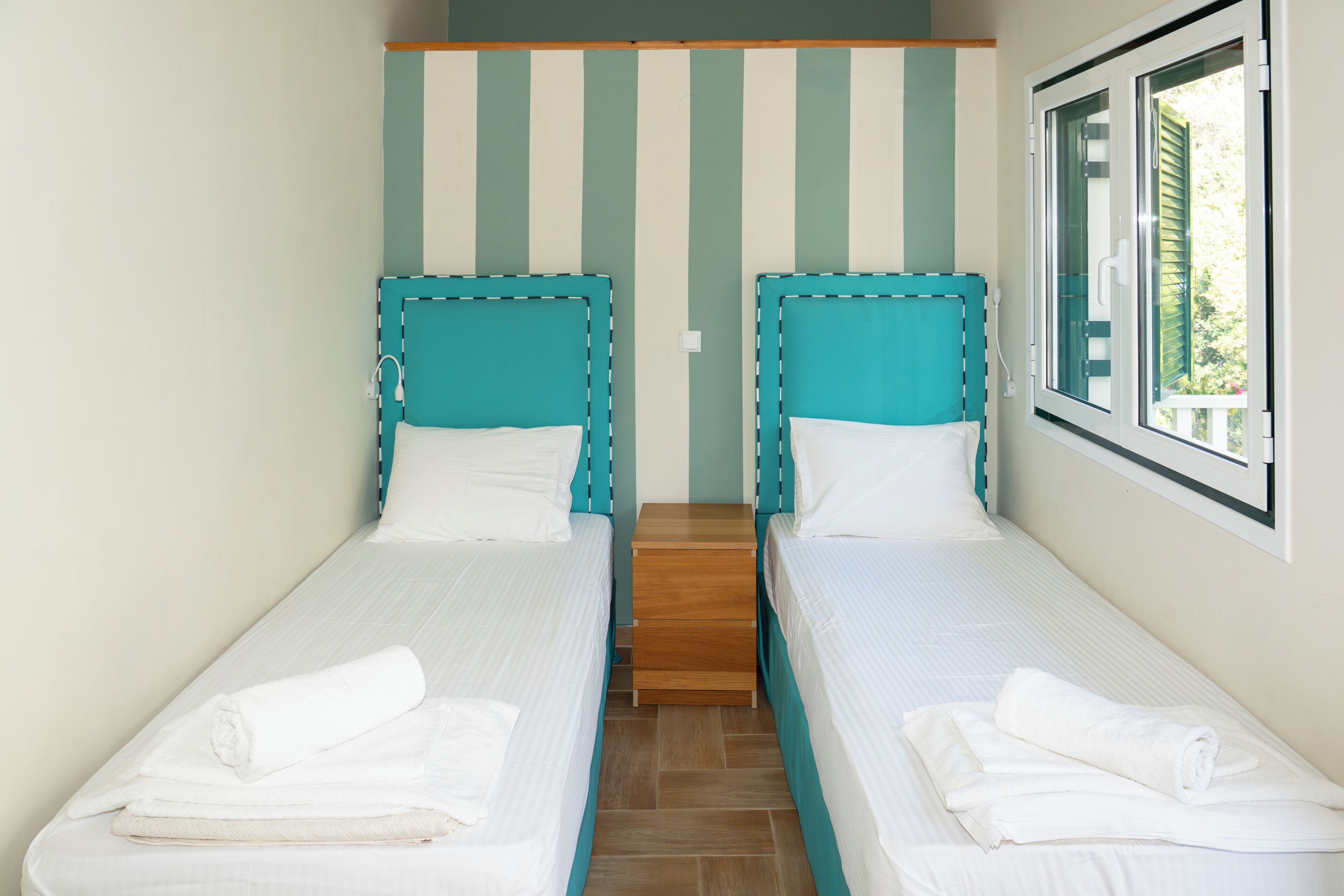 A twin room with two single beds, a wooden nightstand, a striped wall, and a window with blinds.