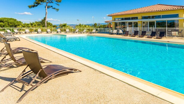 Uma piscina com espreguiçadeiras, um edifício com telhado de azulejos e uma árvore ao fundo.