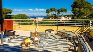 Terraço com mesa posta para refeição, garrafa de vinho e copos, com vista para a costa, árvores e edifícios.