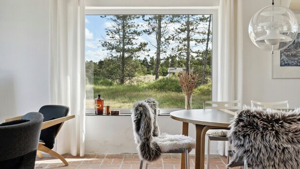 A room with a view of trees, a dining table, chairs, a fur-covered chair, and a hanging light fixture. - Stone house's simplicity in Jerup's vicinity (Jerup)