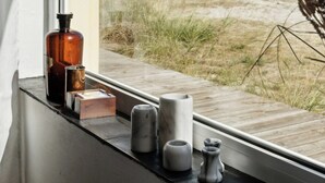 A window with a view of a beach and dunes, a wooden deck, and various decorative items on the windowsill. - Stone house's simplicity in Jerup's vicinity (Jerup)