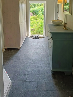 Un pasillo con suelo de piedra que conduce a una puerta con vista a un jardín, un lavabo blanco sobre un mostrador y un espejo encima.
