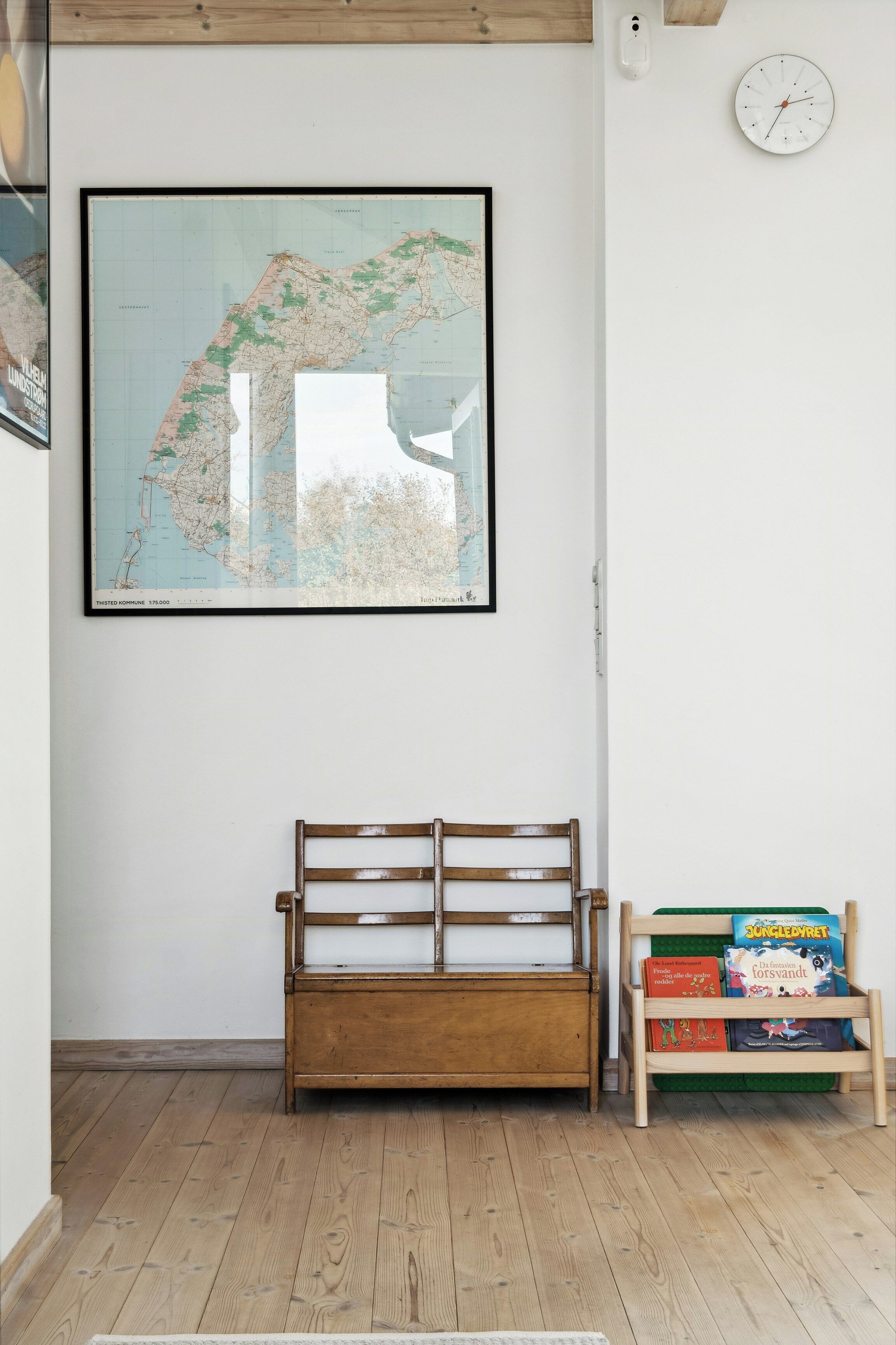 A room with a wooden bench, a bookshelf with books, a framed map, and a wall clock.