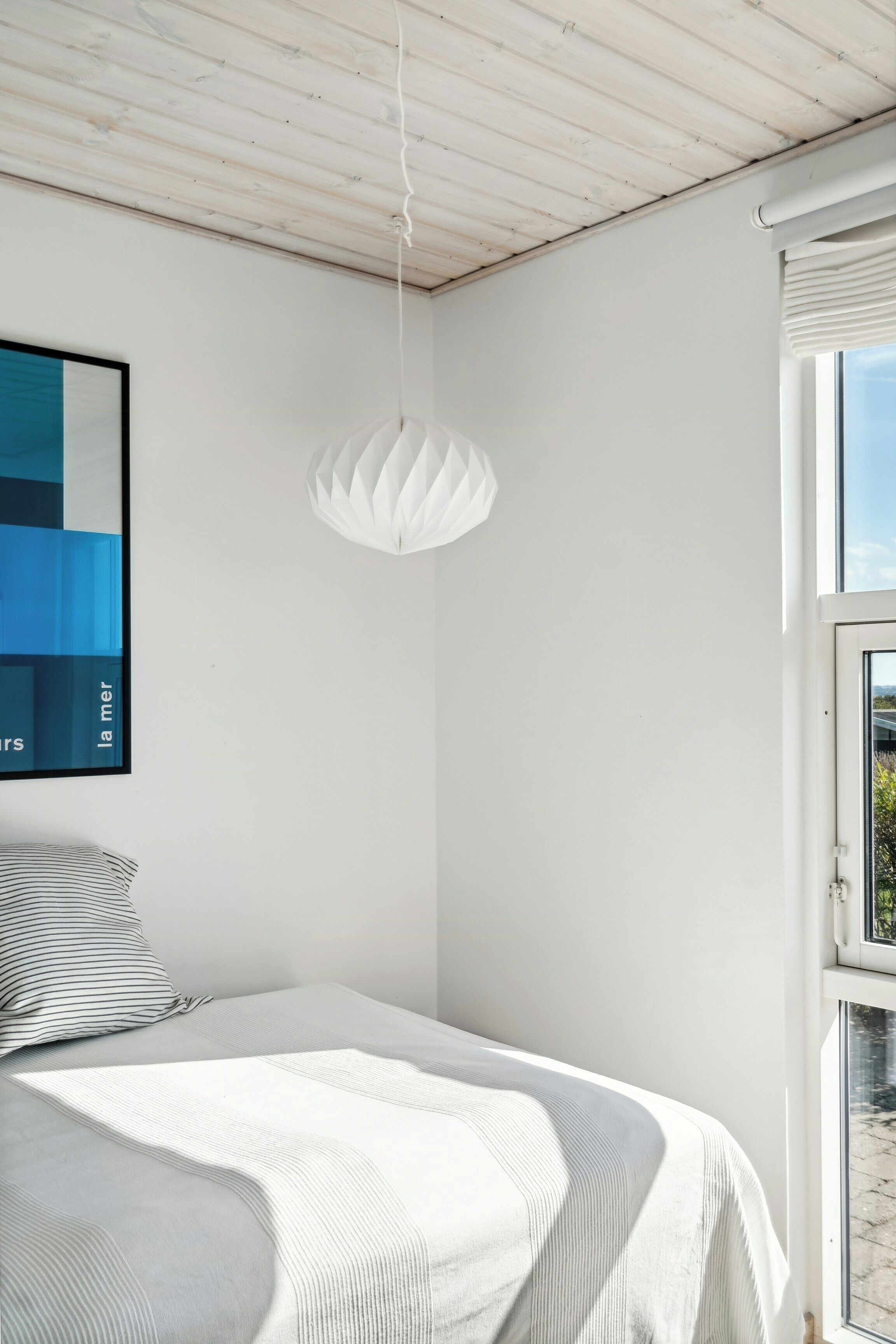 Un dormitorio con una cama, una lámpara colgante, un ventanal y un cuadro abstracto en la pared.