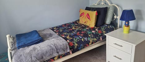 Uma cama de solteiro com colcha florida, manta azul e um xale de tricô cinza. Tem uma mesinha de cabeceira branca com uma luminária e uma gaveta.