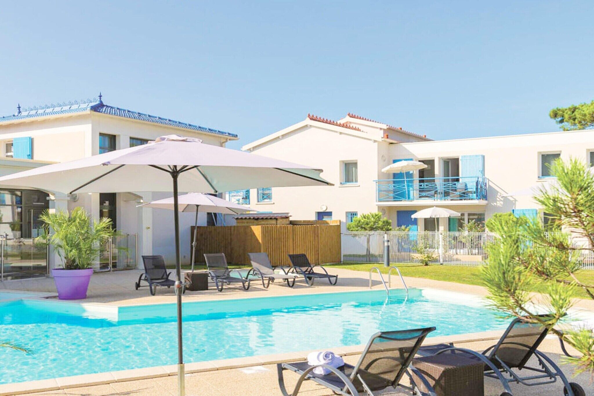 A swimming pool with lounge chairs and umbrellas, surrounded by buildings with balconies and a wooden fence.