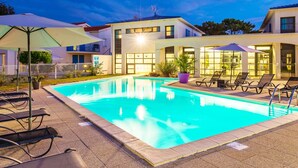 Una piscina con sillones y sombrillas, rodeada de edificios con grandes ventanales y una turbina eólica visible.