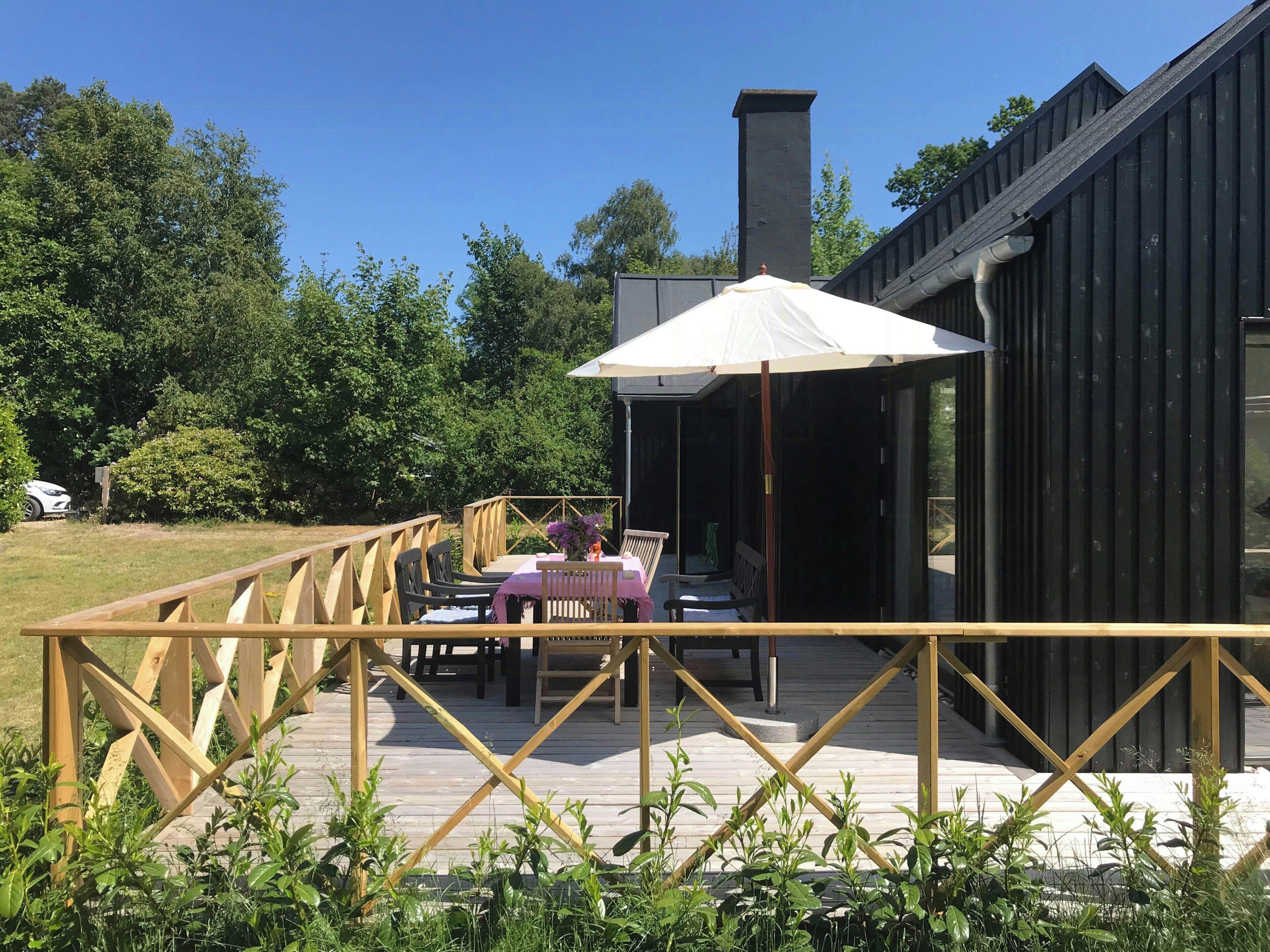 Una terraza de madera con una mesa y sillas, un espacio cubierto con un parasol blanco, y un edificio negro con una chimenea.