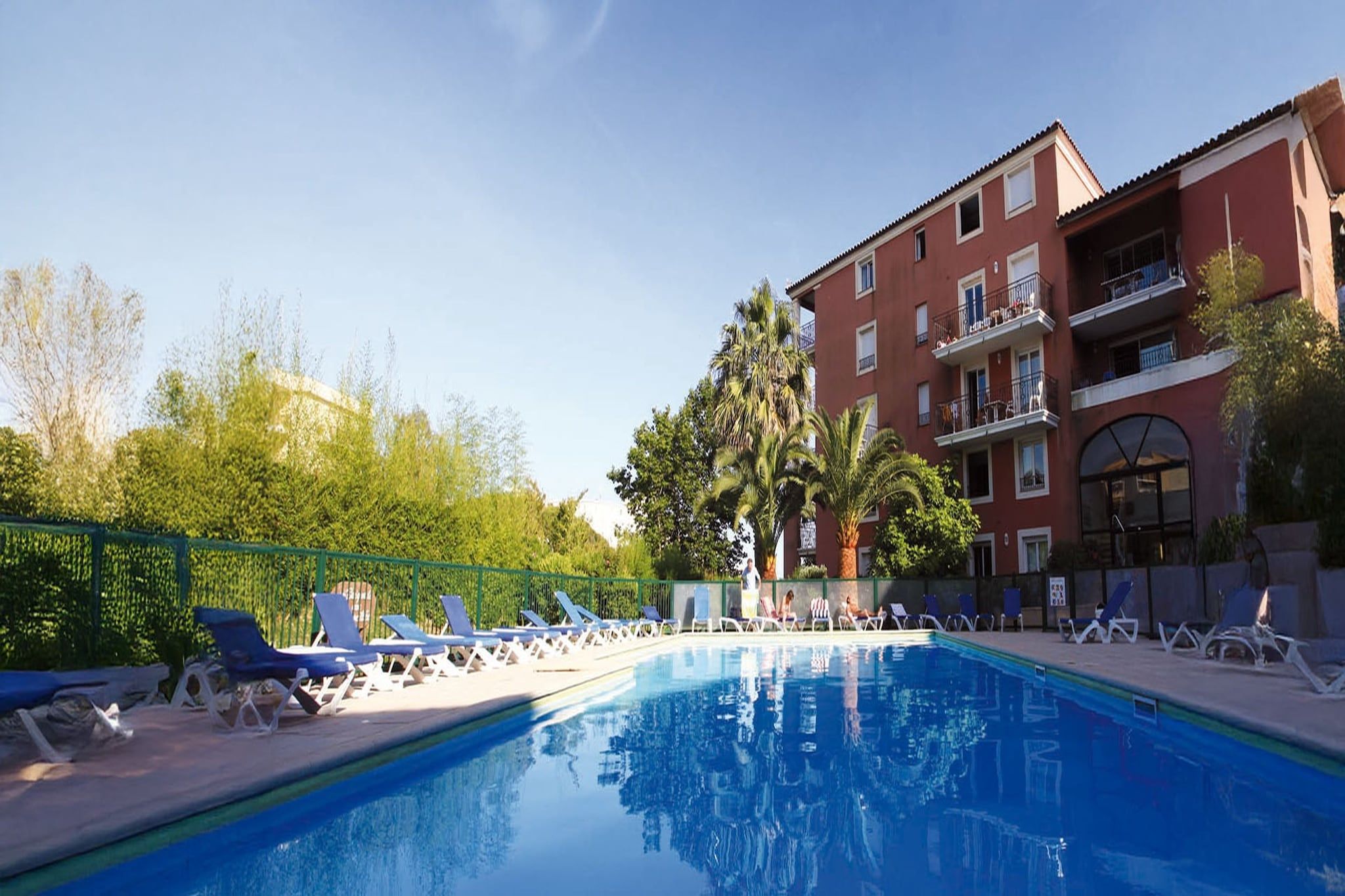 Piscina com espreguiçadeiras, um edifício com varandas e vegetação ao fundo.