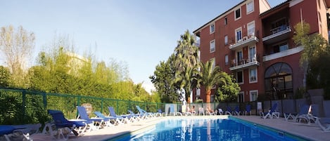 Piscina com espreguiçadeiras, um edifício com varandas e vegetação ao fundo.